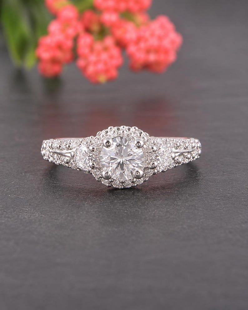 May include: A silver-toned ring featuring a large round center stone surrounded by a halo of smaller stones. The band is split and encrusted with additional small round stones, with two prominent side stones flanking the central halo setting.
