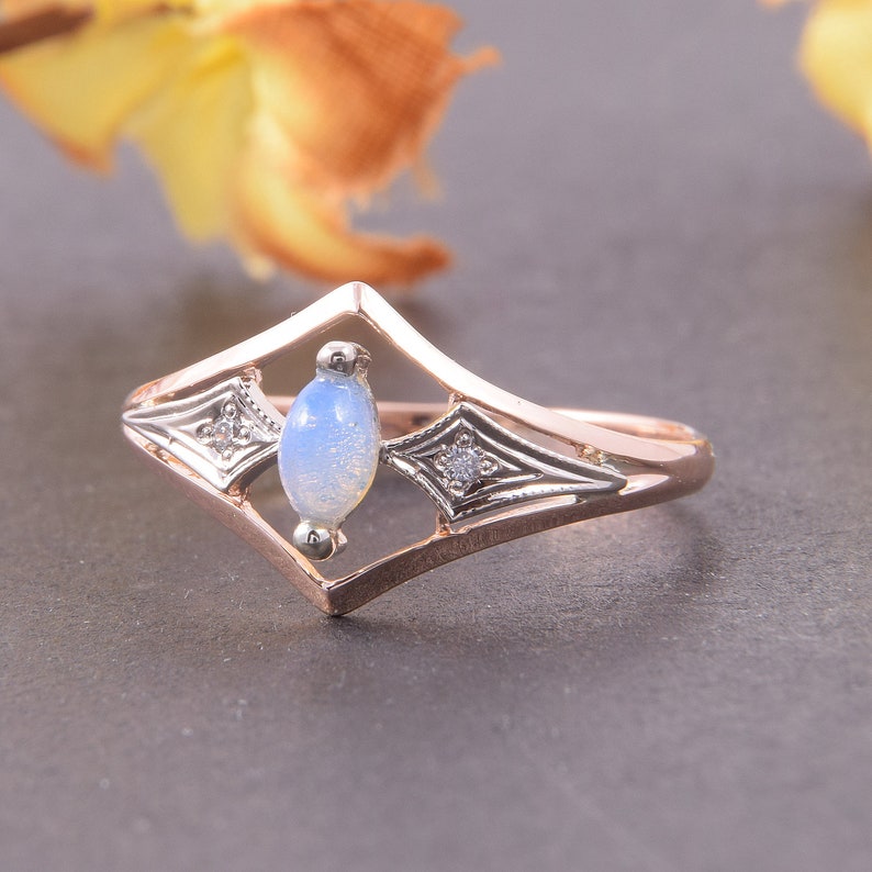 May include: A rose gold ring featuring a central marquise-shaped opal cabochon set within a diamond-shaped frame. The frame includes two small clear accent stones on either side of the opal, set against silver-toned metal detailing.