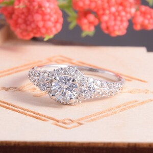 May include: A silver-toned ring featuring a large, round, clear center stone surrounded by a halo of smaller clear stones. The band is split and encrusted with additional small clear stones on both sides, creating a multi-stone design.