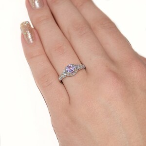 May include: A silver-toned ring featuring a round, light purple center stone set in a band adorned with small, clear, sparkling accent stones. The metal band has a textured, intricate design that extends along the sides of the central setting.