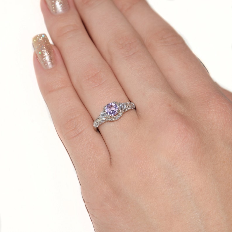 May include: A silver-toned ring featuring a round, light purple center stone set in a band adorned with small, clear, sparkling accent stones. The metal band has a textured, intricate design that extends along the sides of the central setting.