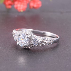 May include: A silver-toned ring featuring a large round-cut central stone surrounded by a halo of smaller stones. Two additional round stones are set on either side, with a band encrusted with multiple tiny clear stones along the sides.