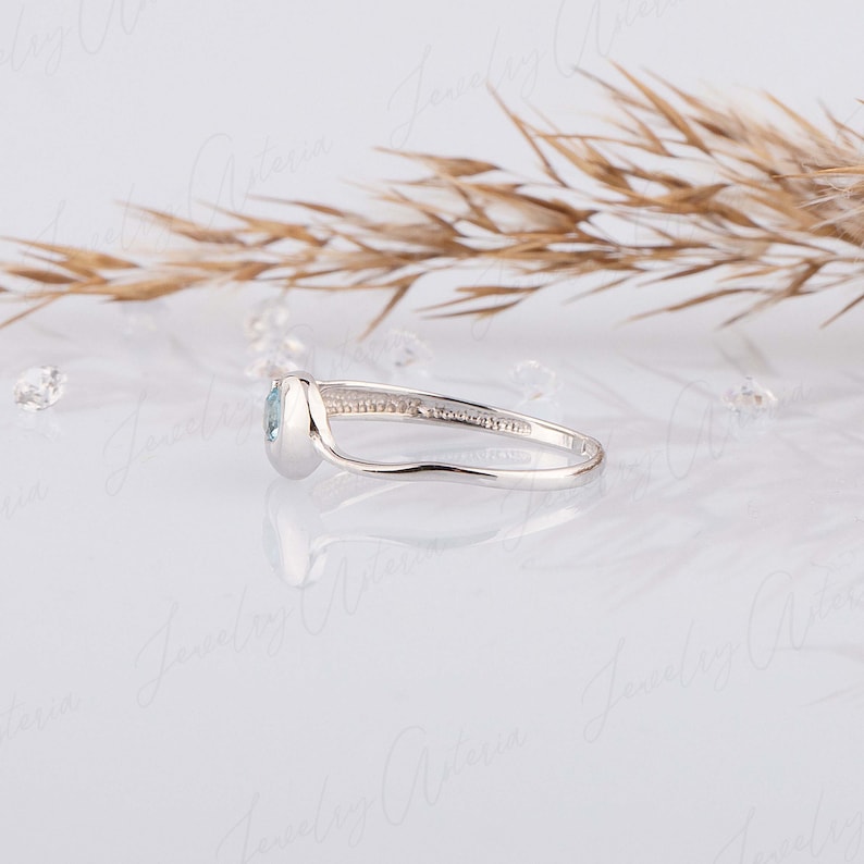 Puede incluir: Un delicado anillo de plata con una piedra preciosa azul claro engastada en garras. El anillo tiene una banda curva y se muestra sobre un fondo blanco.