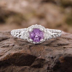 May include: A silver-toned ring featuring a round, faceted purple center stone surrounded by a halo of small clear stones. Two additional clear stones are set on either side of the center, with a band encrusted with small clear stones.