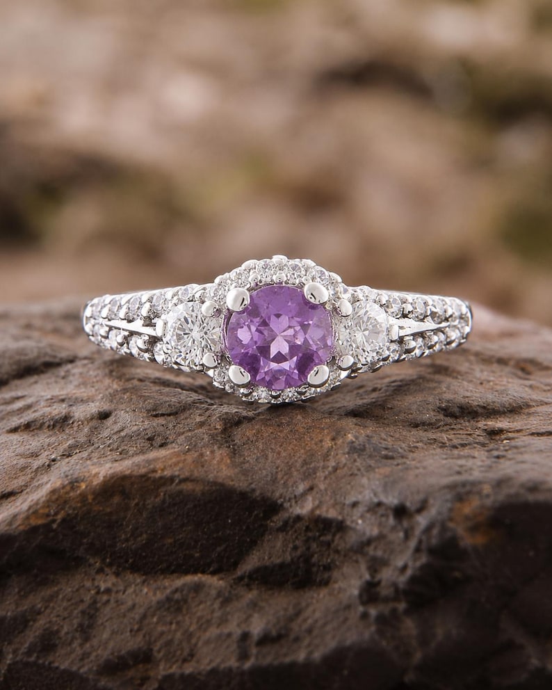 May include: A silver-toned ring featuring a round, faceted purple center stone surrounded by a halo of small clear stones. Two additional clear stones are set on either side of the center, with a band encrusted with small clear stones.