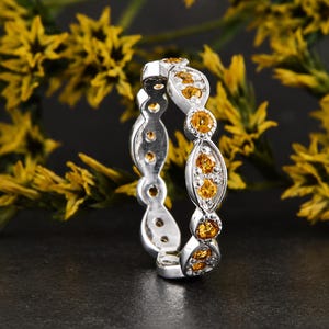May include: A silver eternity band featuring a series of marquise-shaped settings, each containing a round, vibrant yellow citrine gemstone. The metal band has a polished finish, and the stones are arranged in a repeating pattern around the ring.