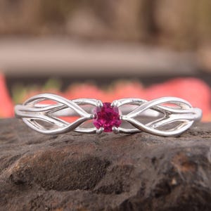 May include: A silver-toned ring featuring a central round-cut vibrant pink gemstone. The band is crafted with an intricate, open-work design that branches out from the center stone, creating a delicate, symmetrical pattern along the finger.