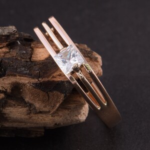 May include: A gold ring with a single, clear, square-shaped gemstone set in the center. The ring has three bands that are intertwined.