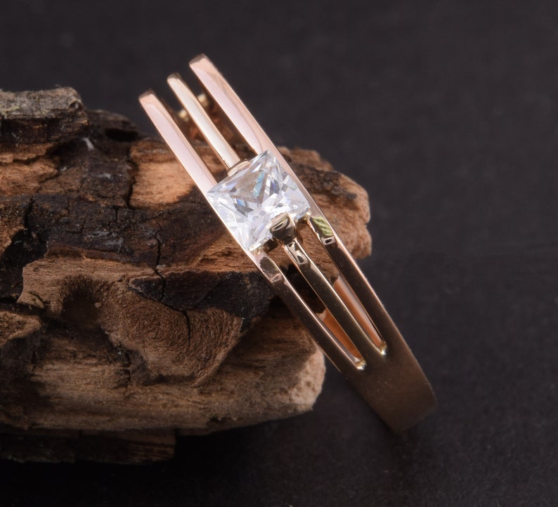 May include: A gold ring with a single, clear, square-shaped gemstone set in the center. The ring has three bands that are intertwined.