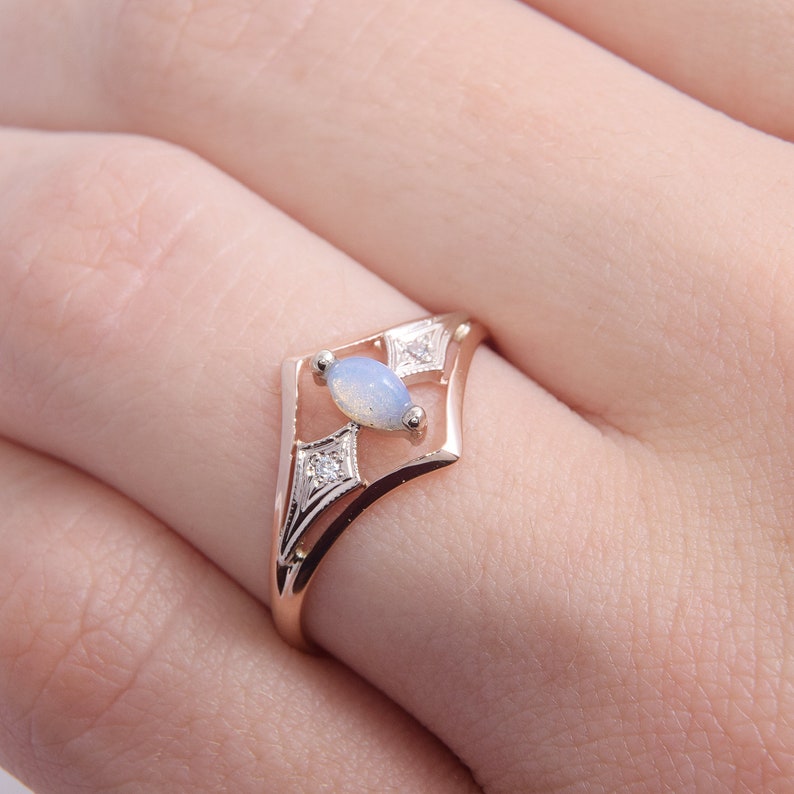 May include: A rose gold ring featuring a central marquise-cut opal stone with a soft blue iridescence. The band has a geometric, open-frame design accented by two small, clear, round-cut diamonds set within silver-toned, diamond-shaped settings.