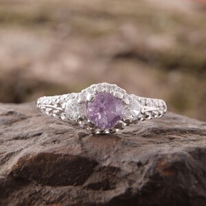 May include: A silver-toned ring featuring a round, light purple center stone surrounded by a halo of small clear stones. Two larger clear stones are set on either side of the center, with additional small clear stones embedded along the band.