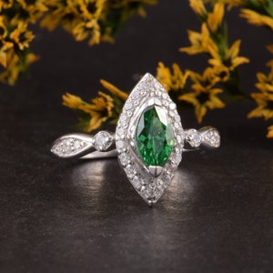 Pode incluir: Um anel de tom prateado com uma pedra preciosa verde central de corte marquise rodeada por um halo de pequenas pedras claras. A banda &eacute; decorada com pedras claras adicionais nas laterais, criando um design detalhado e sim&eacute;trico.