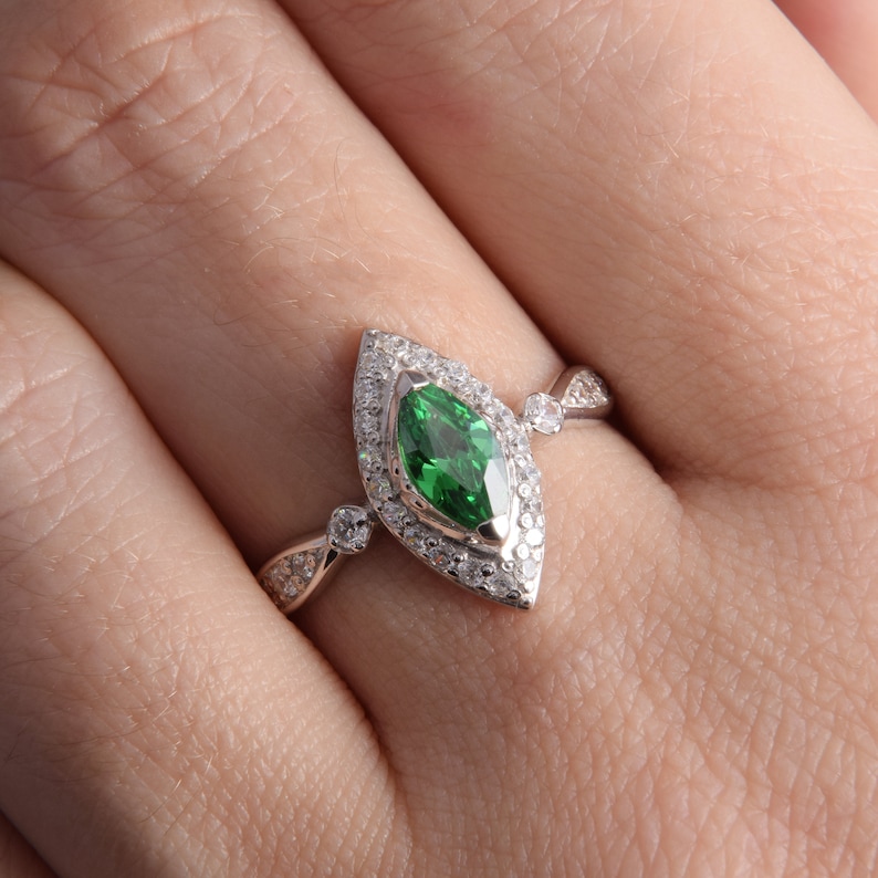Pode incluir: Um anel de tom prateado com uma pedra preciosa verde central de corte marquise rodeada por um halo de pequenas pedras claras. A banda &eacute; acentuada com pedras claras adicionais nas laterais, criando um design detalhado e sim&eacute;trico.