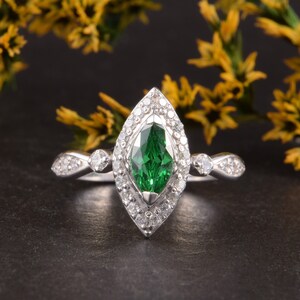 Pode incluir: Um anel de tom prateado com uma pedra preciosa verde central de corte marquise rodeada por um halo de pequenas pedras claras. A banda &eacute; acentuada com pedras claras adicionais em cada lado, criando um design sim&eacute;trico e pontiagudo.