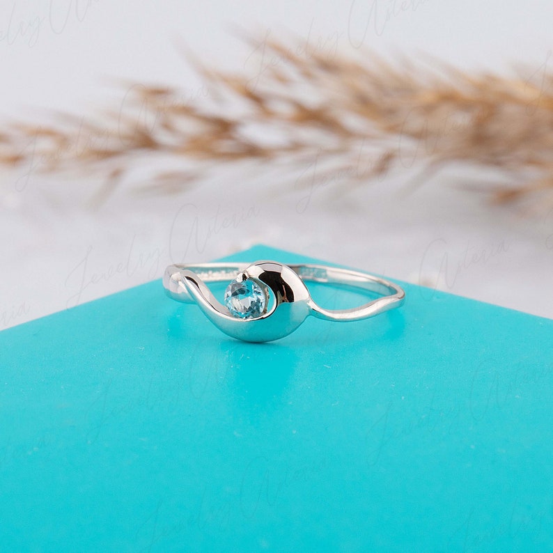 Puede incluir: Un anillo de plata con una piedra preciosa azul claro engastada en una banda con forma de ola.