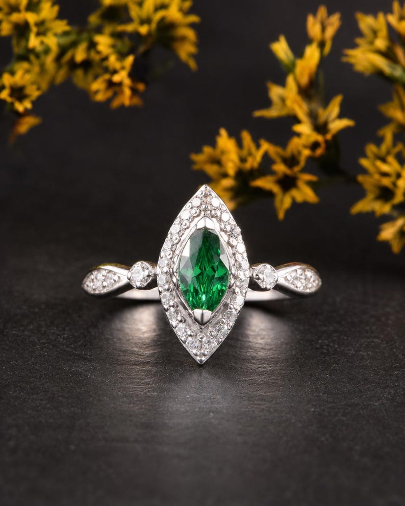 Pode incluir: Um anel de tom prateado com uma pedra preciosa verde central de corte marquise, rodeada por um halo de pequenas pedras claras. A banda inclui pedras claras adicionais em cada lado, criando um design detalhado e sim&eacute;trico.