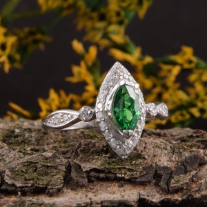 Pode incluir: Um anel de tom prateado com uma pedra preciosa verde central de corte marquise rodeada por um halo de pequenas pedras claras. A banda &eacute; decorada com pedras claras adicionais nas laterais, criando uma apar&ecirc;ncia detalhada e brilhante.