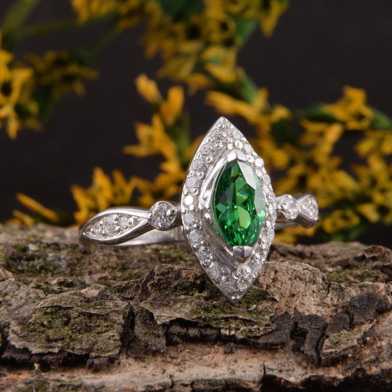 Pode incluir: Um anel de tom prateado com uma pedra preciosa verde central de corte marquise rodeada por um halo de pequenas pedras claras. A banda &eacute; decorada com pedras claras adicionais nas laterais, criando uma apar&ecirc;ncia detalhada e brilhante.