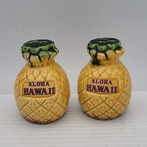 May include: A pair of ceramic salt and pepper shakers shaped like pineapples. Each shaker features a yellow textured body with a green top and the text ALOHA HAWAII printed in red on the front. The shakers have small holes on the top.