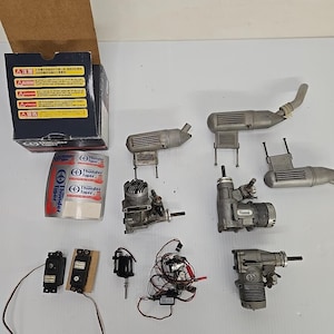 May include: A collection of model airplane engines and associated parts, including silencers, servos, and a control module. A box and tape roll with "Thunder Tiger" branding are also visible. The items are in shades of silver and grey.