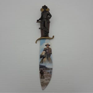 Peut inclure: Un couteau décoratif doté d'un manche métallique sculpté en forme de personnage debout portant un chapeau de cowboy. La lame présente une illustration imprimée colorée d'un cowboy à cheval et d'une silhouette dans un paysage désertique.