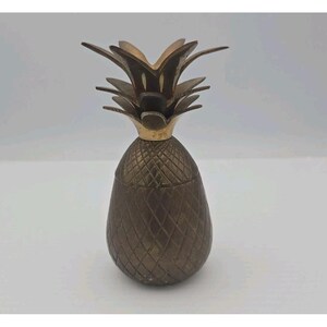 Puede incluir: Un recipiente vintage de latón con forma de piña. El recipiente tiene un cuerpo texturizado con un patrón de diamantes y una corona detallada con hojas. La piña es de un color dorado antiguo cálido.