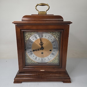 May include: A wooden mantel clock with a dark brown finish, featuring a brass handle on top. The clock face has a gold center, silver chapter ring with black Roman numerals, and ornate gold corner spandrels. The word Howard Miller is visible.