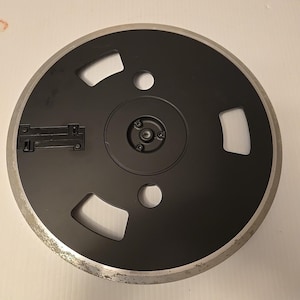 May include: A circular black reel-to-reel tape take-up reel featuring a metallic silver outer rim with visible wear. The center hub is black with a small circular opening, surrounded by three additional cutouts and a rectangular latch mechanism.