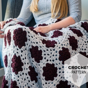 May include: A crochet blanket with a granny square pattern in shades of gray and burgundy. The blanket is draped over a chair. CROCHET PATTERN is written in white text on a white hexagon.