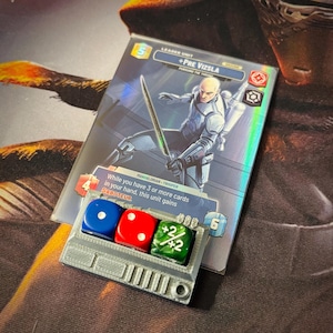 May include: A close-up of a grey plastic dice tray with three dice. The dice are a blue die with a white dot, a red die with a white dot, and a green die with a white plus sign and the number 2 on each side. The dice tray has a slot for each die and a small handle on the side. The dice tray is sitting on top of a card game card with the text "+PRE VIZSLA" and "MANGALORIAN - TROOPER".