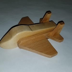 May include: A wooden toy airplane with a light brown finish. The plane has a rounded nose and two vertical stabilizers on the tail.