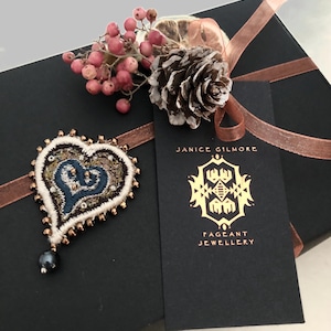 Op de afbeelding: Een zwarte cadeaudoos met een bruin satijnen lint eromheen gebonden. De doos is versierd met een kleine dennenappel en een takje rode bessen. Een hartvormige geborduurde broche met een parel hanger is aan het lint bevestigd. De doos heeft een label dat eraan vastzit met de tekst "Janice Gilmore Pageant Jewellery".