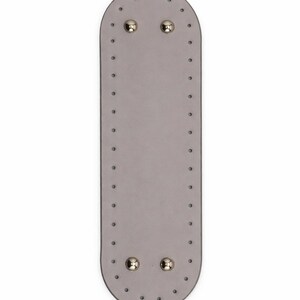 Op de afbeelding: Een langwerpige, ovale, lichtgrijze basis met afgeronde uiteinden, voorzien van een reeks gelijkmatig verdeelde, voorgeponste gaten langs de gehele omtrek en vier gepolijste, zilverkleurige metalen noppen bij de boven- en onderrand.