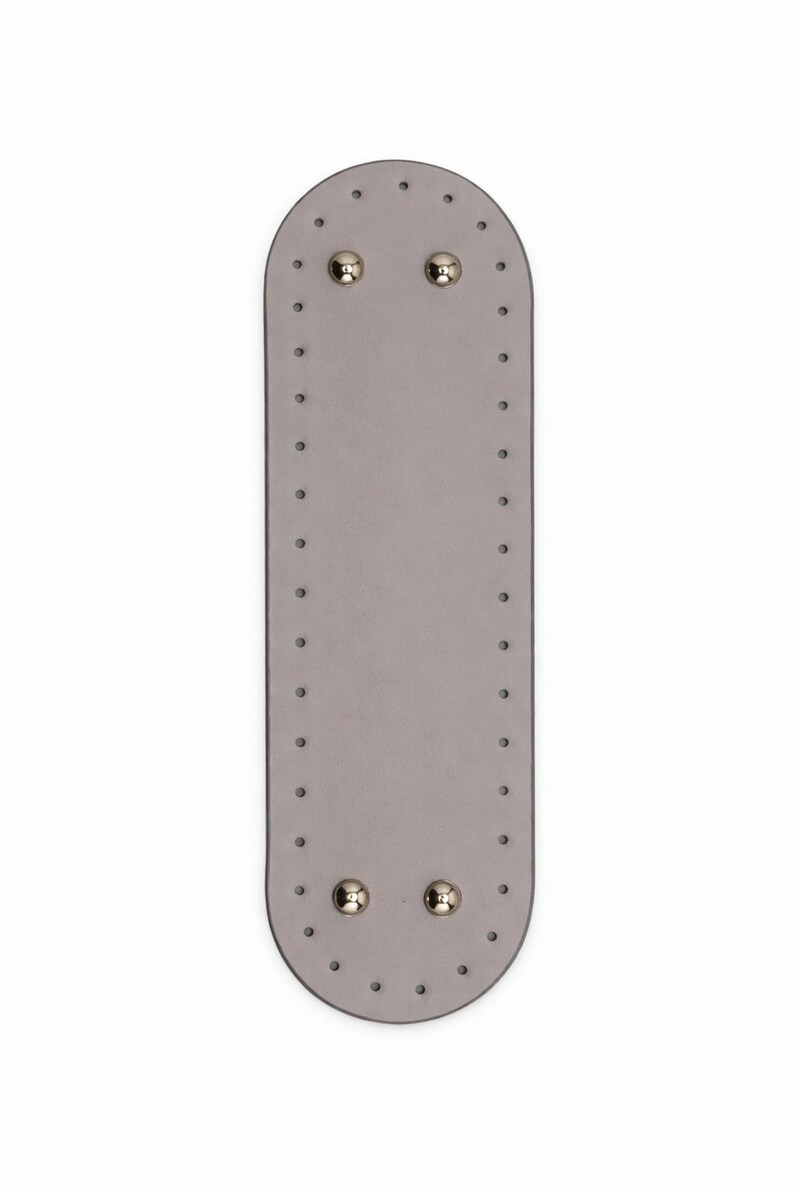 Op de afbeelding: Een langwerpige, ovale, lichtgrijze basis met afgeronde uiteinden, voorzien van een reeks gelijkmatig verdeelde, voorgeponste gaten langs de gehele omtrek en vier gepolijste, zilverkleurige metalen noppen bij de boven- en onderrand.