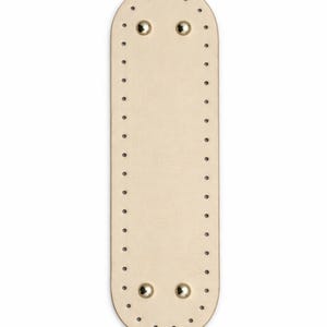 Op de afbeelding: Een langwerpige, ovaalvormige basis van beige leer met afgeronde uiteinden, voorzien van een reeks gelijkmatig verdeelde, voorgeponste gaten langs de gehele rand en vier gepolijste goudkleurige metalen studs bij de boven- en onderkant.