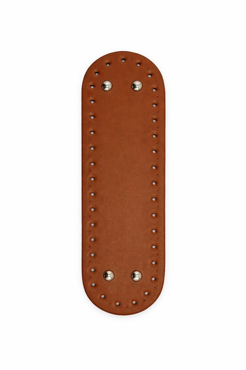 Op de afbeelding: Een langwerpige, ovaalvormige bruine leren patch met een reeks gelijkmatig verdeelde, voorgeponste gaten langs de gehele rand. Vier gepolijste zilverkleurige metalen klinknagels zijn symmetrisch geplaatst aan de boven- en onderkant.