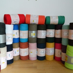 Può includere: Una collezione di 20 bobine di filato di cotone colorato, ciascuna con un'etichetta bianca con il nome del marchio "Lilly" e una piccola illustrazione di una pecora. Il filato è disposto in file su una superficie di legno.