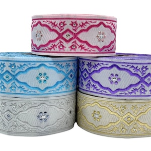 Può includere: Cinque rotoli di nastro decorativo in vari colori, tra cui rosa, blu, viola, grigio e oro. Ogni nastro presenta un design ricamato con un motivo floreale centrale e abbellimenti scintillanti. I nastri sono impilati uno sopra l'altro.