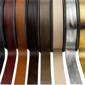 Pode incluir: Uma coleção de rolos de fita de couro em várias cores: preto, bordô, castanho, castanho escuro, bege, prata e dourado. As fitas estão ligeiramente desenroladas, mostrando sua textura e brilho. Ideal para projetos de artesanato e design.