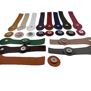 Könnte beinhalten: Eine Sammlung mehrfarbiger Lederriemen mit silberfarbenen Metalldruckknöpfen. Das Set enthält verschiedene Längen und Formen, wobei einige Riemen kreisförmige Enden und andere längliche Designs in Braun-, Weiß-, Rot-, Blau-, Grün- und Grautönen aufweisen.