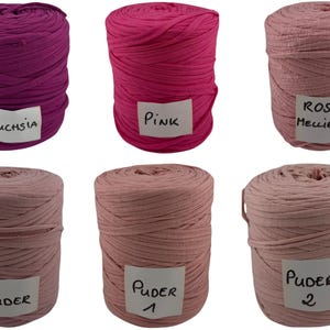 Könnte beinhalten: Sechs zylindrische Garnrollen aus Stoff in zwei Reihen angeordnet. Die obere Reihe zeigt fuchsiafarbenes, pinkes und hellrosa Garn mit den Etiketten Fuchsia, Pink und Rosa Melliert. Die untere Reihe zeigt drei Rollen in Altrosa mit den Etiketten Puder, Puder 1 und Puder 2.