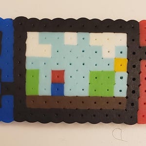 May include: A pixel art representation of a video game console, crafted from colorful plastic beads. The console features a black screen displaying a pixelated landscape with blue, green, and red elements. The controllers are blue and red.