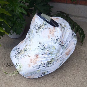 Puede incluir: Una funda blanca para asiento de coche de beb&eacute; con un estampado floral en tonos verdes, grises y marrones. La funda est&aacute; colocada sobre un asiento de coche, con el asa visible. El dise&ntilde;o floral incluye hojas y otros elementos bot&aacute;nicos.