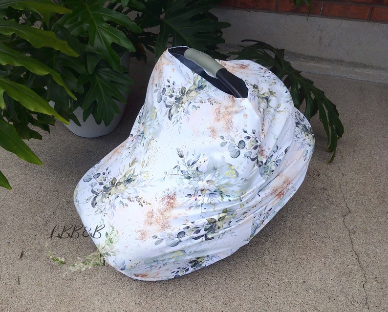 Puede incluir: Una funda blanca para asiento de coche de beb&eacute; con un estampado floral en tonos verdes, grises y marrones. La funda est&aacute; colocada sobre un asiento de coche, con el asa visible. El dise&ntilde;o floral incluye hojas y otros elementos bot&aacute;nicos.