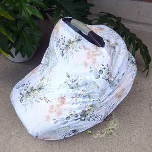 Puede incluir: Una funda blanca para asiento de coche de beb&eacute; con un dise&ntilde;o floral con hojas verdes y azules y detalles dorados. La funda est&aacute; colocada sobre un asiento de coche, proporcionando sombra y protecci&oacute;n. El estampado floral a&ntilde;ade un toque de elegancia.