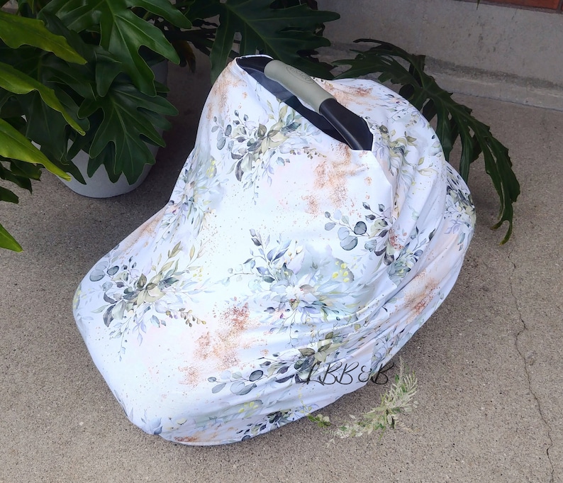 Puede incluir: Una funda blanca para asiento de coche de beb&eacute; con un dise&ntilde;o floral con hojas verdes y azules y detalles dorados. La funda est&aacute; colocada sobre un asiento de coche, proporcionando sombra y protecci&oacute;n. El estampado floral a&ntilde;ade un toque de elegancia.