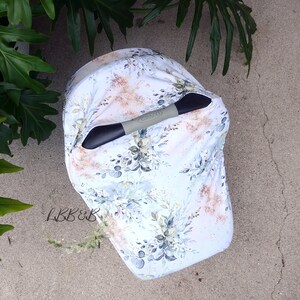 Puede incluir: Una funda blanca para asiento de coche de beb&eacute; con un dise&ntilde;o floral en tonos azules, verdes y dorados. La funda est&aacute; colocada sobre un asa negra y gris con la marca "Graco". El texto "LBB&B" est&aacute; en la esquina inferior izquierda.