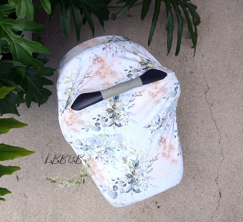Puede incluir: Una funda blanca para asiento de coche de beb&eacute; con un dise&ntilde;o floral en tonos azules, verdes y dorados. La funda est&aacute; colocada sobre un asa negra y gris con la marca "Graco". El texto "LBB&B" est&aacute; en la esquina inferior izquierda.