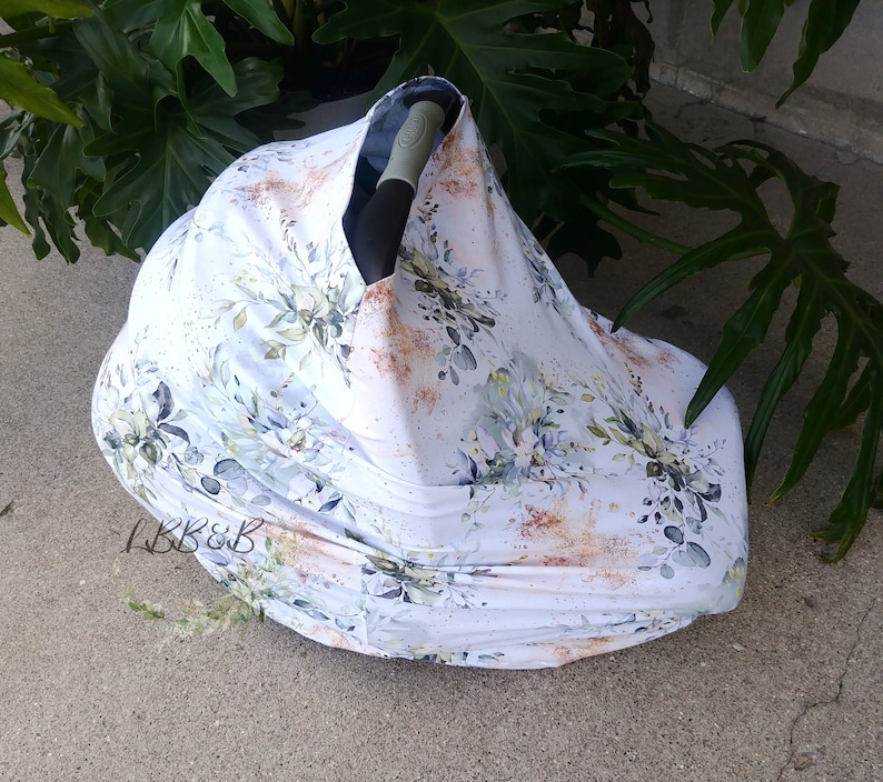 Puede incluir: Una funda blanca para asiento de coche de beb&eacute; con un estampado floral en tonos verdes, azules y marrones. La funda est&aacute; colocada sobre un asiento de coche, con el asa visible. El texto "LBB&B" est&aacute; en la esquina inferior izquierda.