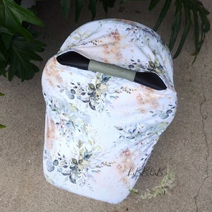 Puede incluir: Una funda blanca para asiento de coche de beb&eacute; con un dise&ntilde;o floral en tonos verdes, grises y dorados. La funda tiene un asa Graco y est&aacute; colocada sobre una superficie de hormig&oacute;n. El patr&oacute;n floral incluye hojas y ramas.